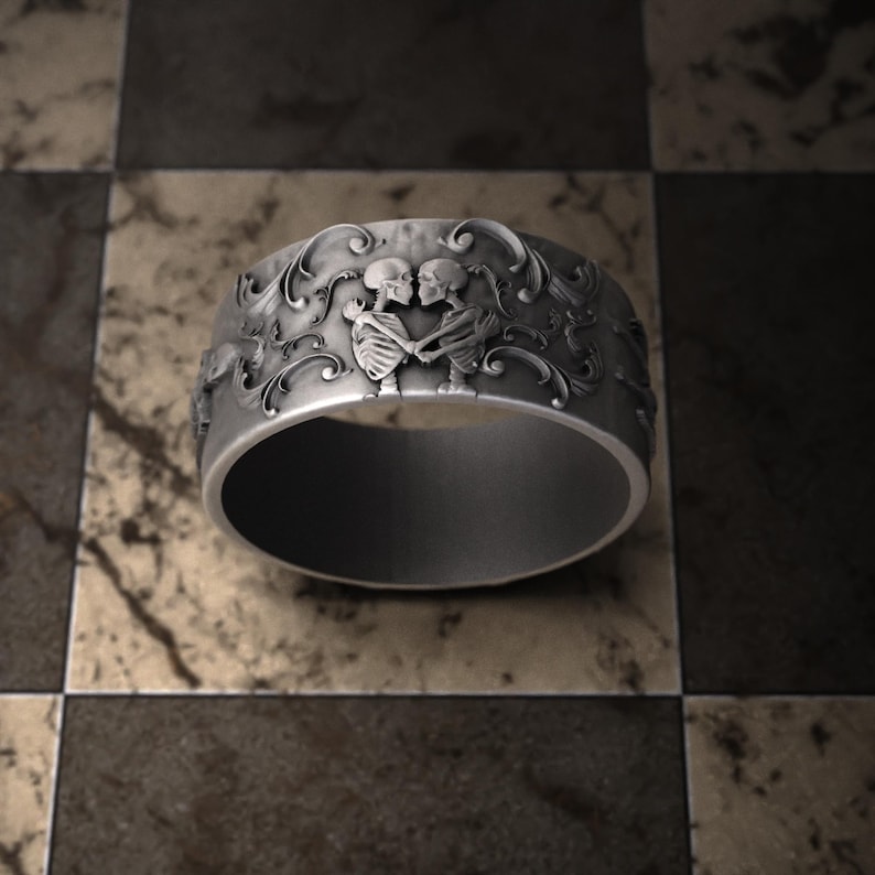Pu&ograve; includere: Un anello color argento con un design dettagliato raffigurante due scheletri che si abbracciano, circondati da motivi floreali ornati. L'anello ha una fascia larga e una finitura lucida, adatta a uno stile gotico o alternativo.