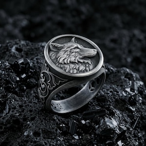 May include: A silver-toned ring featuring a circular face with a detailed relief of a wolf head in profile. The band displays intricate scrollwork patterns on the sides and includes the hallmark 925 stamped on the inner surface of the metal.