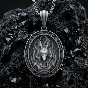 Könnte beinhalten: Ein silberfarbener, ovaler Anhänger mit einer Reliefdarstellung der ägyptischen Gottheit Anubis, umgeben von stilisierten Flammen. Der Anhänger ist von einem Mäander-Muster umrandet und hängt an einer passenden silberfarbenen Kette aus Vierkantgliedern.