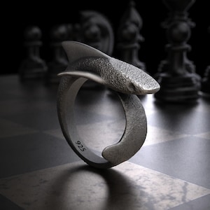 May include: A silver ring with a shark design. The ring features a textured shark body and fin that wraps around the finger. The number "925" is stamped on the ring. The ring is displayed on a chessboard.