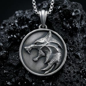 May include: A circular silver-toned pendant featuring a raised, detailed relief of a snarling wolf head in profile. The pendant has a brushed metal finish and is suspended from a matching silver-toned box chain link necklace.