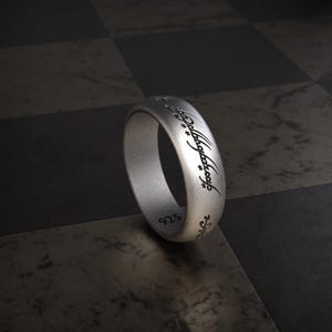 Könnte beinhalten: Ein silberner Ring mit eingravierter Schrift. Der Ring hat eine glatte, matte Oberfläche und die Zahl "925" ist sichtbar. Die Schrift ist in einer stilisierten Schriftart gehalten. Der Ring ist ein Schmuckstück.