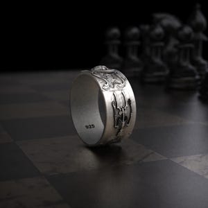 Pu&ograve; includere: Un anello d'argento con un design dettagliato raffigurante uno scheletro e motivi ornati. L'anello &egrave; contrassegnato con "925" ed &egrave; posto su uno sfondo scuro con pezzi degli scacchi.