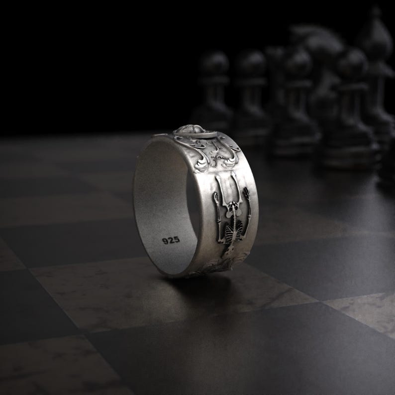 Pu&ograve; includere: Un anello d'argento con un design dettagliato raffigurante uno scheletro e motivi ornati. L'anello &egrave; contrassegnato con "925" ed &egrave; posto su uno sfondo scuro con pezzi degli scacchi.