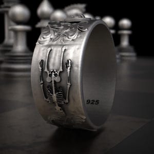 Pu&ograve; includere: Un anello d'argento con un design dettagliato a scheletro. L'anello presenta una figura di scheletro in rilievo con le braccia tese e decorazioni a volute. La fascia &egrave; larga e ha il numero "925" stampato. L'anello &egrave; esposto su una superficie scura.