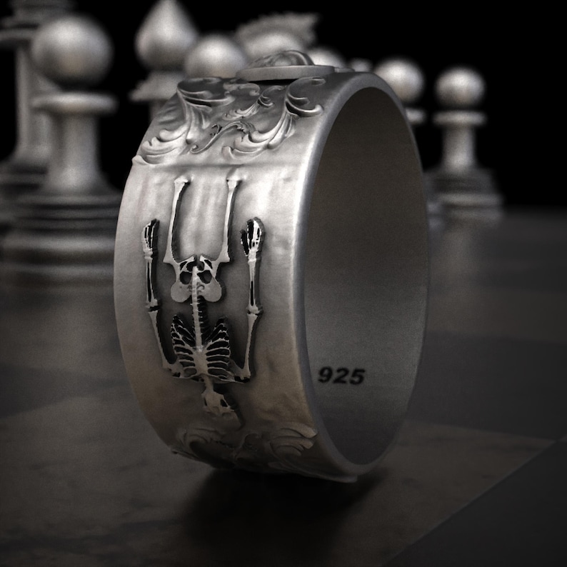 Pu&ograve; includere: Un anello d'argento con un design dettagliato a scheletro. L'anello presenta una figura di scheletro in rilievo con le braccia tese e decorazioni a volute. La fascia &egrave; larga e ha il numero "925" stampato. L'anello &egrave; esposto su una superficie scura.