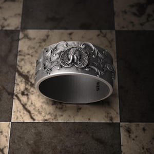 Pu&ograve; includere: Un anello d'argento con un design ornato. L'anello presenta un emblema centrale di un elefante all'interno di una cornice a forma di cuore, circondato da motivi dettagliati e vorticosi. L'anello &egrave; posto su uno sfondo a scacchi.