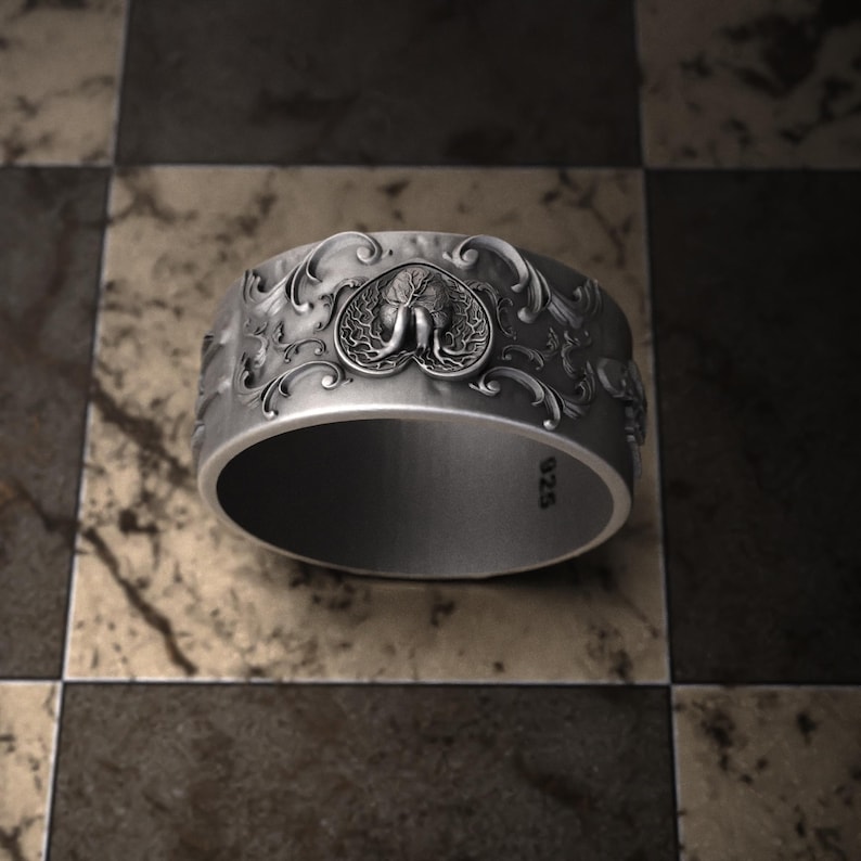Pu&ograve; includere: Un anello d'argento con un design ornato. L'anello presenta un emblema centrale di un elefante all'interno di una cornice a forma di cuore, circondato da motivi dettagliati e vorticosi. L'anello &egrave; posto su uno sfondo a scacchi.