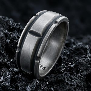 May include: A wide, brushed silver-tone ring with a textured, industrial aesthetic. The band features a raised central section with evenly spaced vertical notches along the edges. The interior of the band is clearly stamped with the number 925.