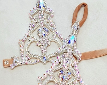 May include: A pair of white foot jewelry with crystal embellishments. The jewelry is attached to a brown strap.