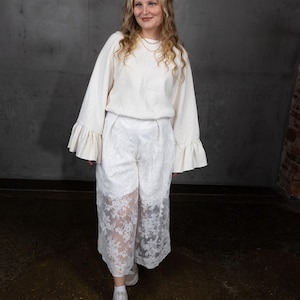 Könnte beinhalten: Cremefarbenes Outfit mit einem langärmeligen Oberteil mit Rüschenärmeln und einer Hose mit weitem Bein und Spitzendetails. Das Outfit wird mit weißen Turnschuhen kombiniert. Der Hintergrund ist eine graue Wand und ein dunkler Boden.