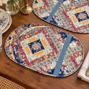 May include: Two oval quilted placemats featuring a patchwork design with floral and geometric patterns in shades of red, blue, yellow, and cream. The placemats have a stitched texture and a blue fabric border, showcasing a central diamond motif.
