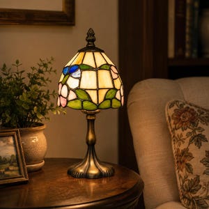 May include: A stained glass table lamp with a dome-shaped shade featuring cream, green, blue, and pink glass panels arranged in a floral pattern. The lamp has a dark bronze-toned metal base with a flared foot and a decorative finial on top.