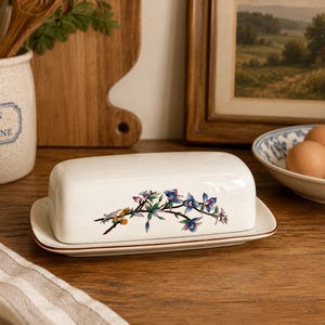 May include: A white ceramic butter dish with a floral design in blue, purple, and yellow. The dish is rectangular with a lid and sits on a matching base. The kitchenware is on a wooden surface.