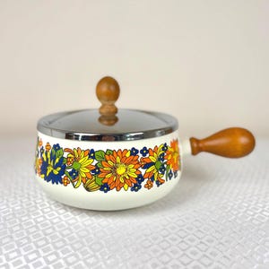 Op de afbeelding: Een vintage witte emaille steelpan met een bloemenpatroon in oranje, geel, blauw en groen. Het heeft een houten handvat en knop, en een roestvrijstalen rand. Een retro keukenartikel.