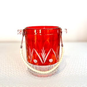 May include: A vibrant red glass ice bucket with a decorative etched design. The bucket has a clear base and a white beaded handle. The handles are attached to the bucket with silver metal. The ice bucket is sitting on a white surface.