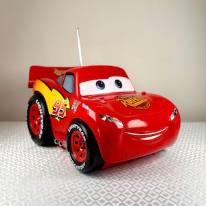 Può includere: Un'auto giocattolo rossa con il numero 95 sul lato e la scritta "LIGHTYEAR" sugli pneumatici. L'auto ha una faccia sorridente e uno spoiler. È un personaggio di Saetta McQueen del film Cars.