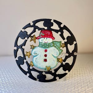 May include: A snowman Christmas ornament. The snowman has a red hat, green scarf, and three red buttons. It is surrounded by gold stars and a black, decorative, circular frame.