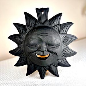May include: A black sun face wall hanging with a smiling expression. The sun has detailed rays and a carved face with closed eyes and a golden-coloured mouth. The wall decor is made of wood and has a hole at the top for hanging.