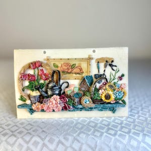 Peut inclure: Sculpture décorative rectangulaire en relief représentant une scène de jardin. Elle comprend un arrosoir, des fleurs, un nichoir et une image florale encadrée. La palette de couleurs comprend du rose, du bleu, du vert et du jaune.