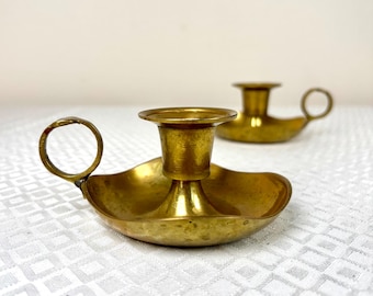May include: Two brass candle holders with a polished metallic finish. Each piece features a wide, shallow, scalloped base, a central cylindrical socket for a candle, and a small, circular finger loop handle attached to the side.