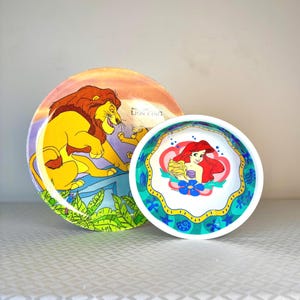 Op de afbeelding: Twee decoratieve borden met Disney-personages. E&eacute;n bord toont Simba uit The Lion King, met een gele leeuw en een zonsondergangachtergrond. Het andere bord toont Ariel uit The Little Mermaid, met een roodharige zeemeermin.
