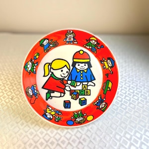 May include: A vintage, round, red and white children's plate. The plate features a central illustration of two cartoon children playing with alphabet blocks. The rim is decorated with cartoon figures. The plate is made of a durable material.