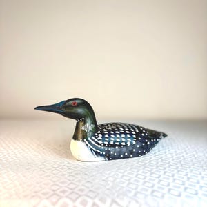 May include: A hand-painted wooden loon figurine. The bird has a black head and back with white dotted patterns, a white belly, and a long black beak. The loon has red eyes and is resting on a white surface.