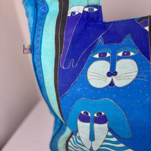 May include: A blue tote bag with a whimsical design featuring stylized animal faces. The bag's main color is a vibrant blue, with accents of light blue, black, and gold. The artwork includes a cat, a dog, and a horse.