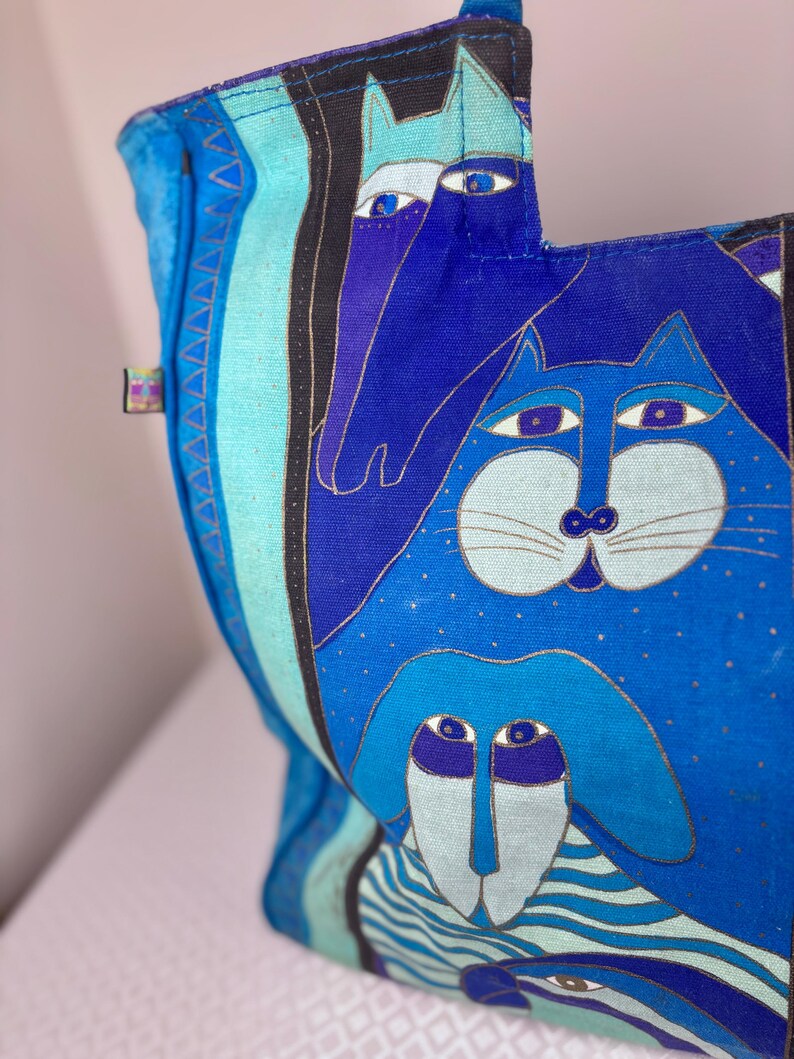 May include: A blue tote bag with a whimsical design featuring stylized animal faces. The bag's main color is a vibrant blue, with accents of light blue, black, and gold. The artwork includes a cat, a dog, and a horse.