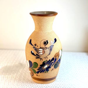 May include: A tan ceramic vase with a bird and floral design. The vase has a brown rim and base, with a painted bird in the centre and blue, green, and white floral accents. The vase has a wide base and a flared top.