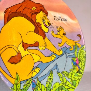 Op de afbeelding: Een ronde decoratieve plaat met een sc&egrave;ne uit Disney's "The Lion King". De afbeelding toont een grote gele leeuw met een bruine manen en een kleinere leeuwenwelp. De achtergrond omvat een berg en weelderig groen gebladerte.