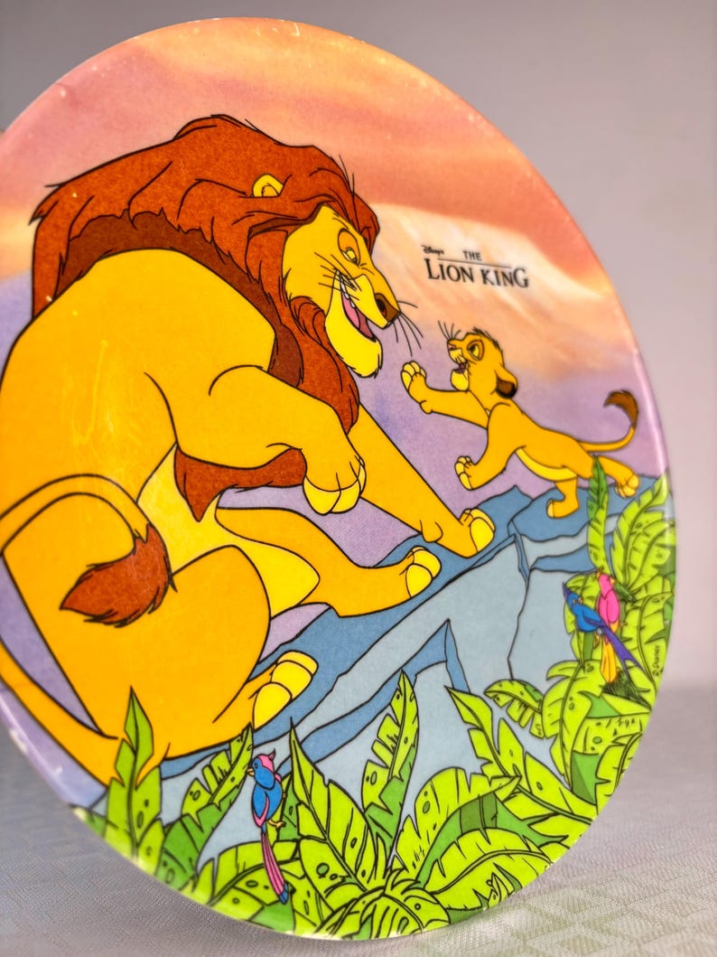 Op de afbeelding: Een ronde decoratieve plaat met een sc&egrave;ne uit Disney's "The Lion King". De afbeelding toont een grote gele leeuw met een bruine manen en een kleinere leeuwenwelp. De achtergrond omvat een berg en weelderig groen gebladerte.
