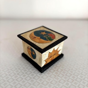 May include: A small, square decorative box with a black base and lid. The box is painted with celestial imagery, including a crescent moon, stars, and a sun with a smiling face. The box has a gold and cream colored background with a crackle finish.