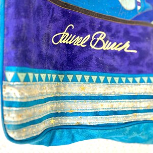 May include: Close-up of a blue tote bag with the name "Laurel Burch" in gold script. The bag features a vibrant design with turquoise, blue, and gold accents. The design includes triangles, stars, and stripes.