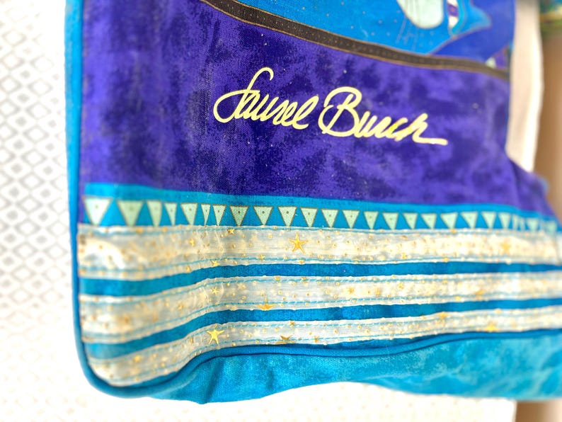 May include: Close-up of a blue tote bag with the name "Laurel Burch" in gold script. The bag features a vibrant design with turquoise, blue, and gold accents. The design includes triangles, stars, and stripes.