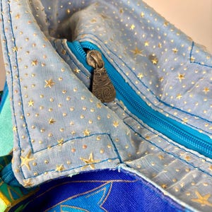May include: Close-up of a light blue fabric bag with a zipper. The bag is decorated with gold stars and dots. The zipper is turquoise, and the zipper pull has a cat design. The bag has a colorful patterned design.