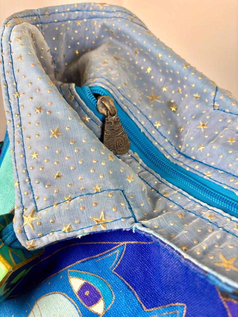May include: Close-up of a light blue fabric bag with a zipper. The bag is decorated with gold stars and dots. The zipper is turquoise, and the zipper pull has a cat design. The bag has a colorful patterned design.