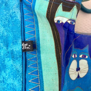 May include: Close-up of a blue tote bag with a cat design. The bag features a vibrant mix of blues and turquoise, with gold accents. The bag has a tag that reads "Gisel Buri".
