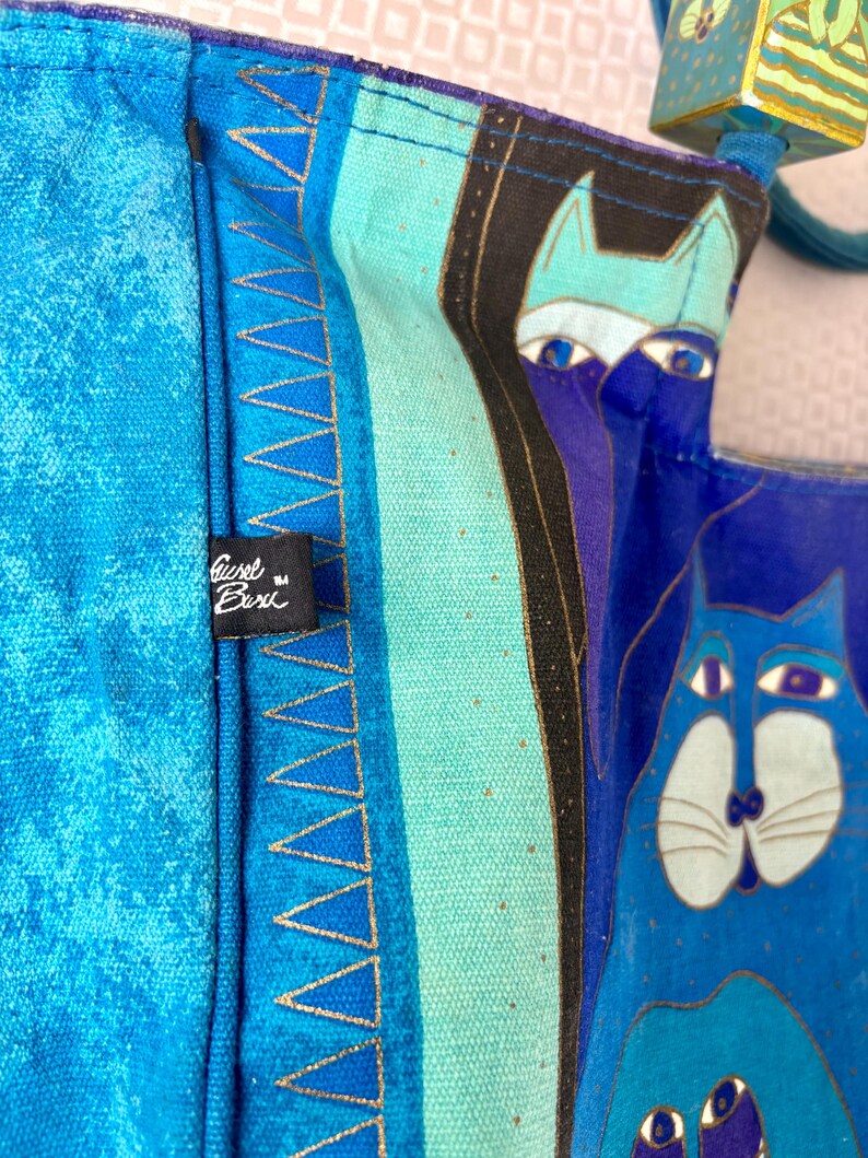 May include: Close-up of a blue tote bag with a cat design. The bag features a vibrant mix of blues and turquoise, with gold accents. The bag has a tag that reads "Gisel Buri".