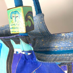 May include: Close-up of a blue tote bag with a colorful design featuring stylized faces and gold accents. The bag has blue handles and a decorative wooden cube with painted images. The interior lining is light blue with gold stars.