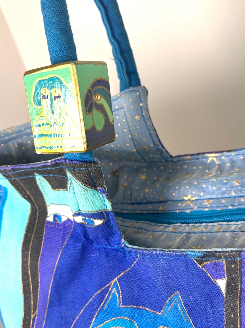 May include: Close-up of a blue tote bag with a colorful design featuring stylized faces and gold accents. The bag has blue handles and a decorative wooden cube with painted images. The interior lining is light blue with gold stars.