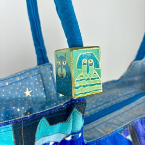 May include: A blue tote bag with a decorative wooden cube featuring painted cat and person designs. The bag has blue handles and a patterned fabric with stars and cat faces. The cube is painted with teal, gold, and blue.