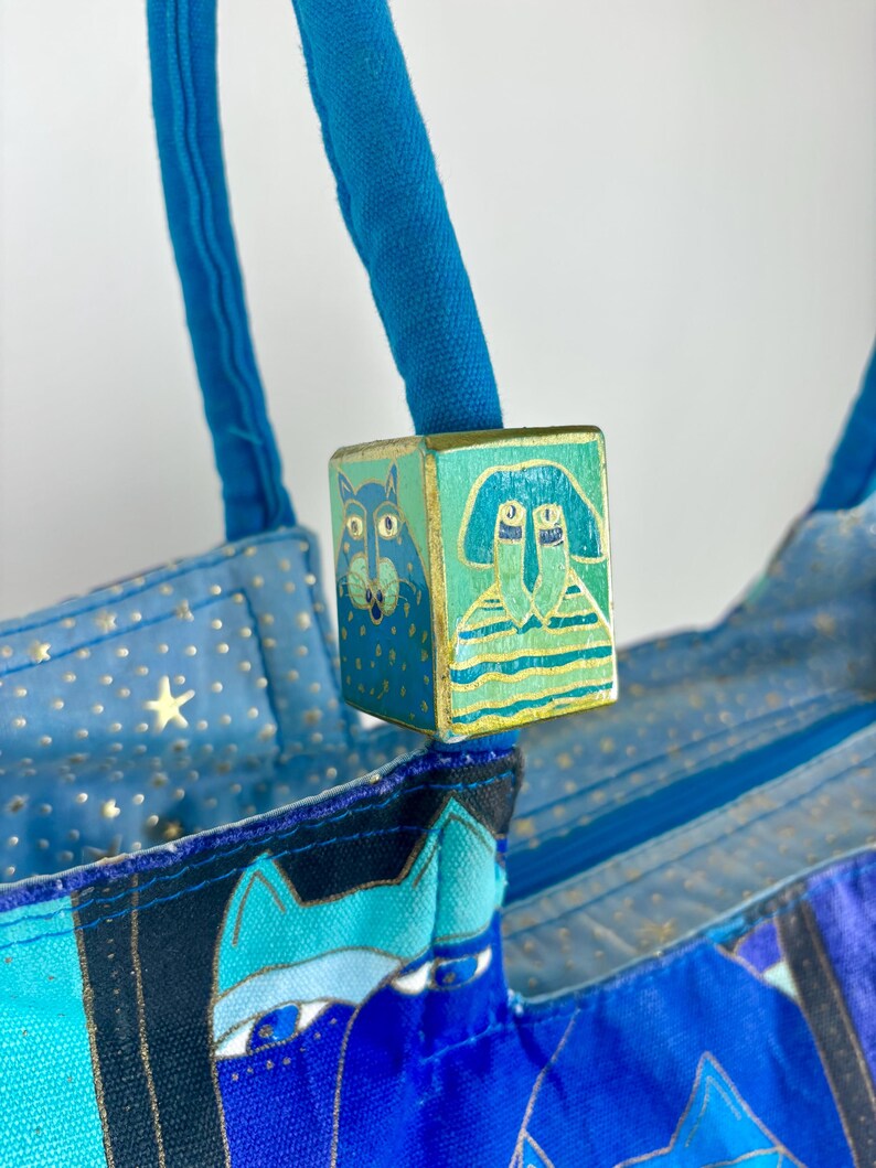 May include: A blue tote bag with a decorative wooden cube featuring painted cat and person designs. The bag has blue handles and a patterned fabric with stars and cat faces. The cube is painted with teal, gold, and blue.