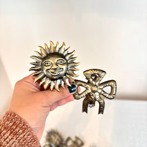 Puede incluir: Pomos de armario en tono bronce con un diseño de sol sonriente y un lazo. El pomo del sol tiene una cara detallada con rayos radiantes. El pomo del lazo tiene detalles intrincados. Se ven pomos adicionales en el fondo.