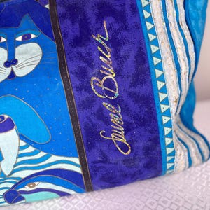 May include: Blue tote bag with a whimsical cat and dog design. The bag features a vibrant color palette of blues, with gold accents and the text "Jewel Burch" in gold glitter. The bag has a striped pattern on the side.
