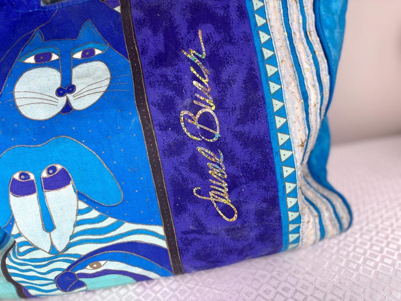 May include: Blue tote bag with a whimsical cat and dog design. The bag features a vibrant color palette of blues, with gold accents and the text "Jewel Burch" in gold glitter. The bag has a striped pattern on the side.