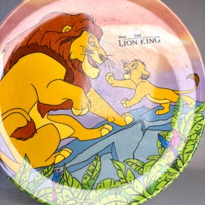 Op de afbeelding: Ronde decoratieve plaat met een sc&egrave;ne uit Disney's The Lion King. De plaat toont Mufasa en Simba met een jungle-achtergrond. De plaat heeft een groene rand met bladeren en een roze en paarse achtergrond.
