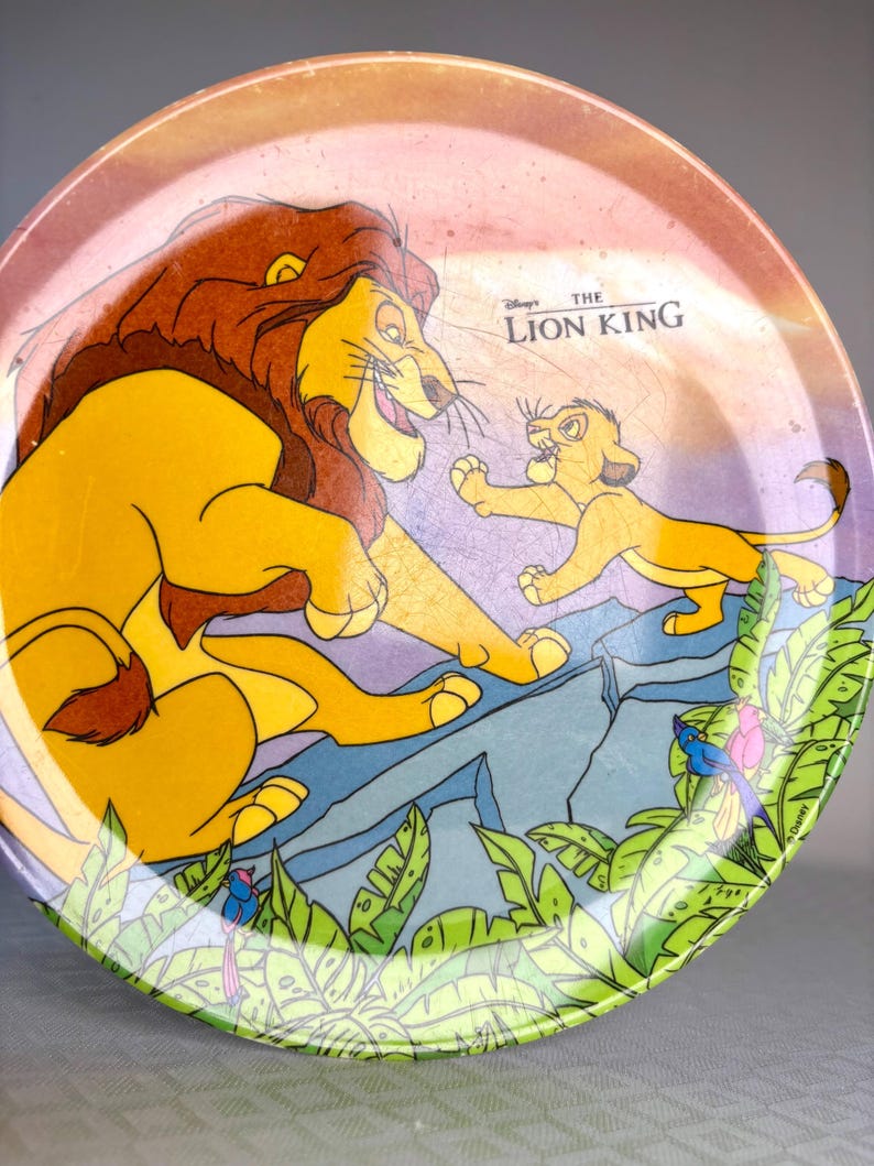Op de afbeelding: Ronde decoratieve plaat met een sc&egrave;ne uit Disney's The Lion King. De plaat toont Mufasa en Simba met een jungle-achtergrond. De plaat heeft een groene rand met bladeren en een roze en paarse achtergrond.