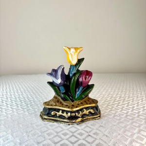 May include: A cast iron tulip figurine featuring yellow, purple, and red flowers. The flowers are set on a black base with gold trim. This decorative piece is a charming accent for any room.